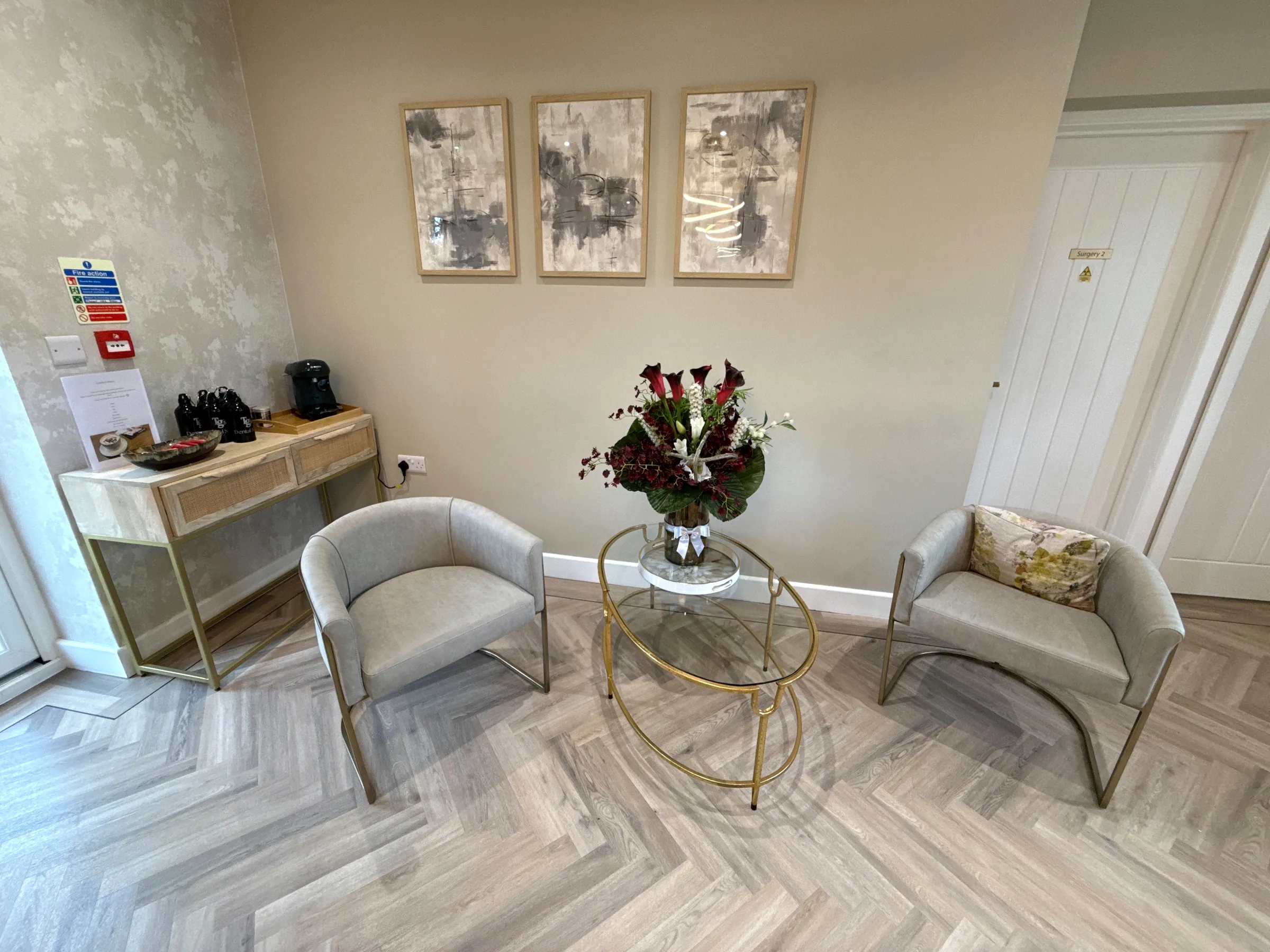 Two upholstered tub chairs and a glass table with a flower arrangement, under three framed abstract prints